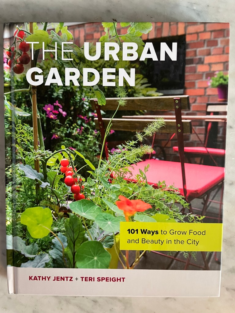 Front cover of a gardening book showing an urban garden with a red chair, red tomatoes and other flowers in front of a brick wall. The Urban Garden book by Kathy Jentz and teri Speight.