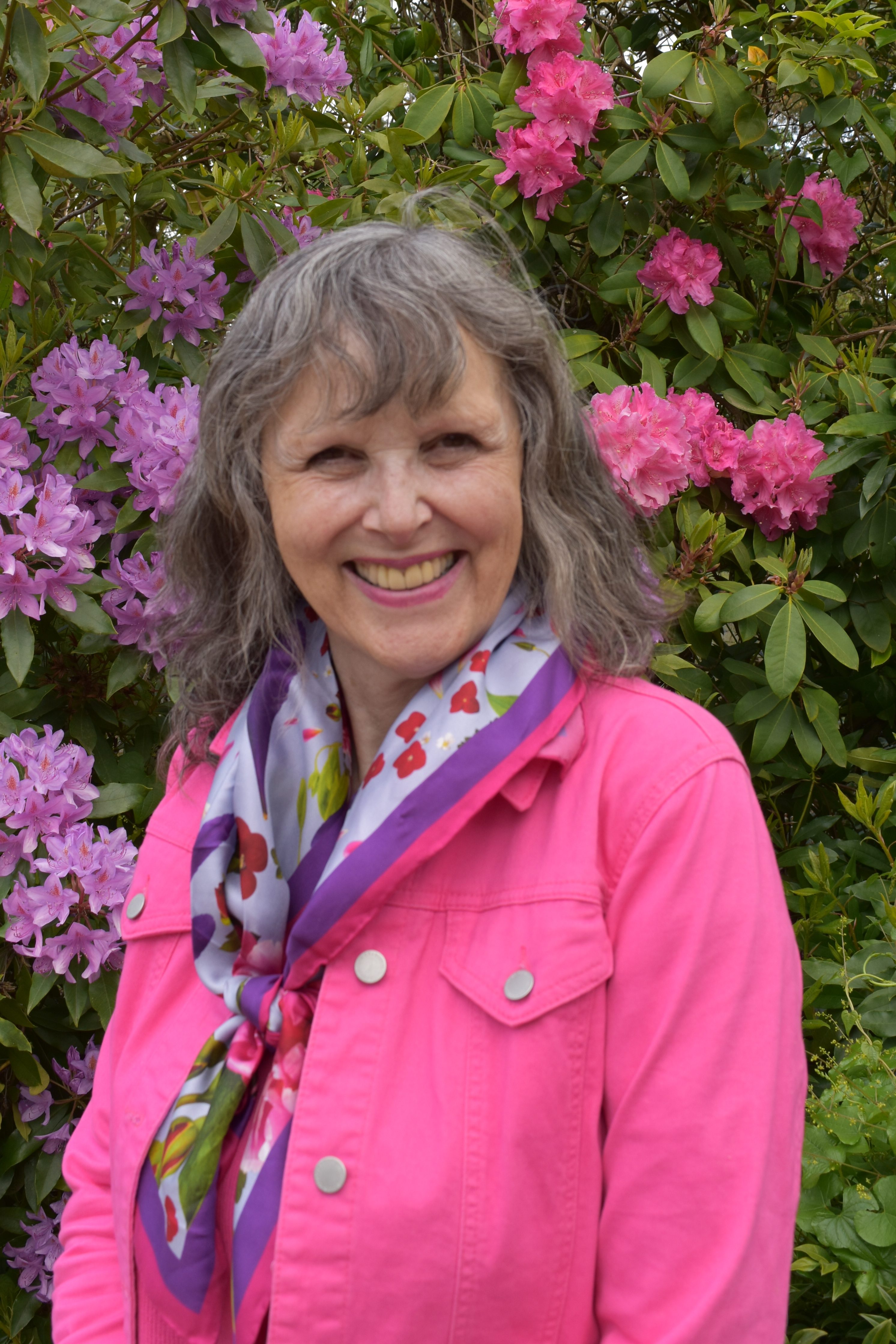 Jenny Rose Carey - garden author in from of pink and purple rhodendron flowers. She is wearing a pink and purple floral scarf and a pink jacket.