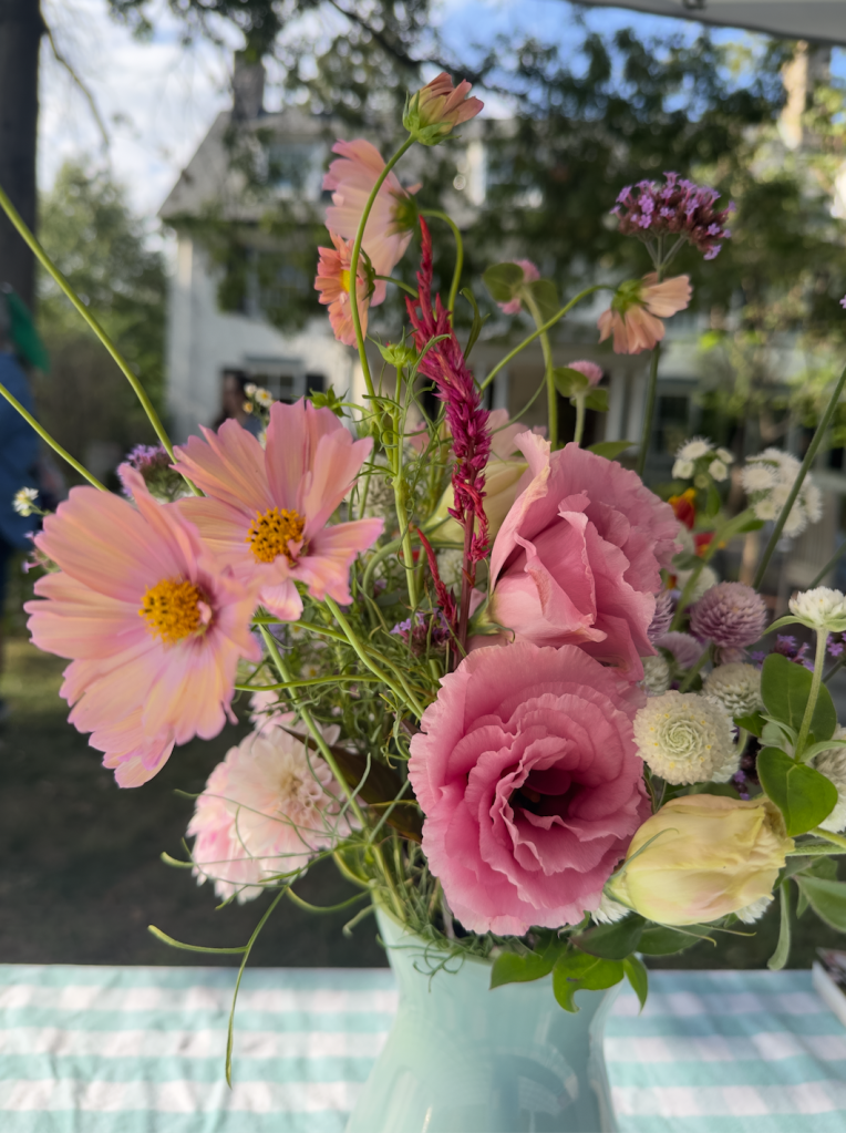 A vase of flowers with cosmos, amaranthus, lisianthus, and more - pink and cream colored on a green and cream checkered tablecloth in front of a house